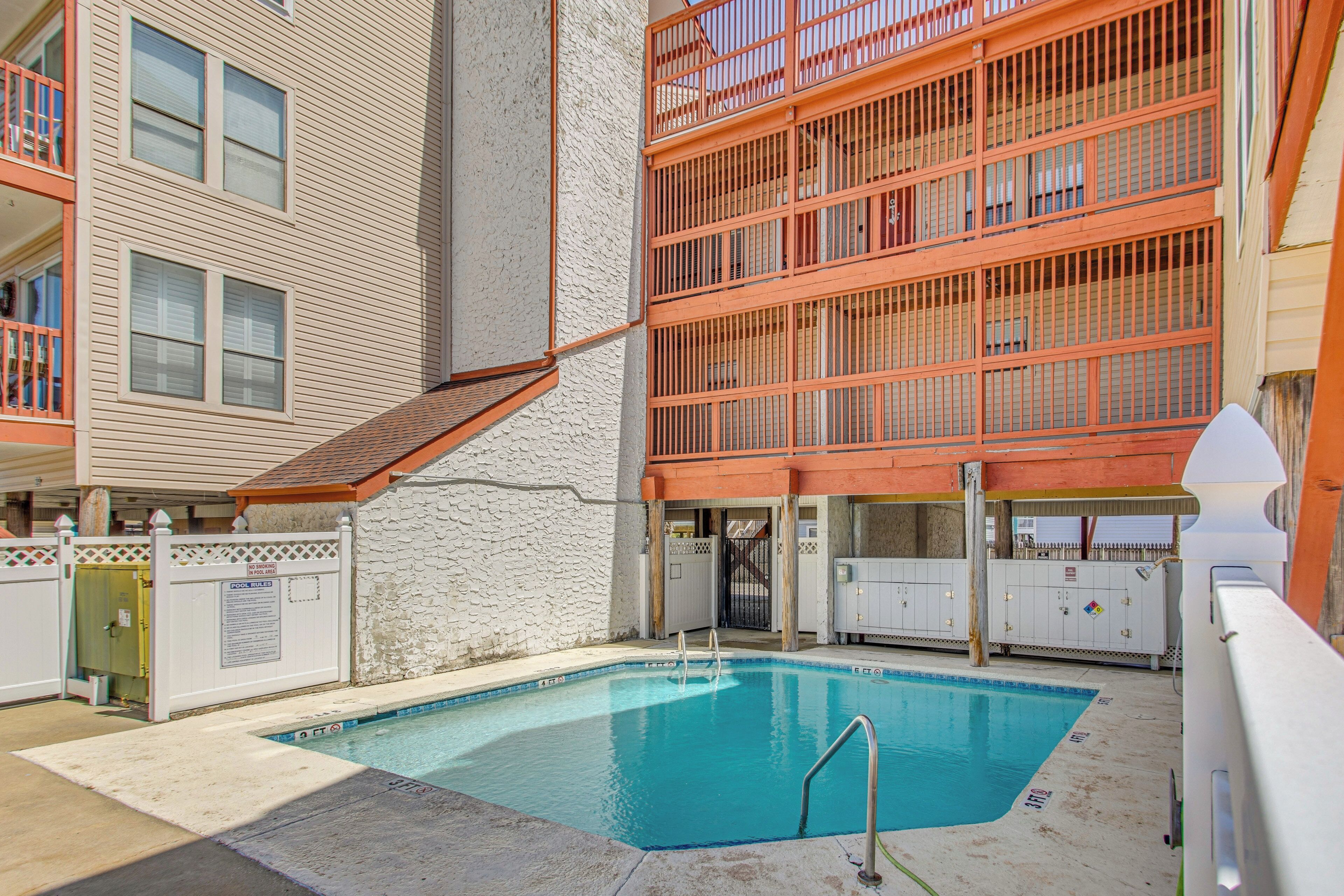 An outdoor swimming pool with a white fence, a stone wall, and a multi-story building with balconies.
