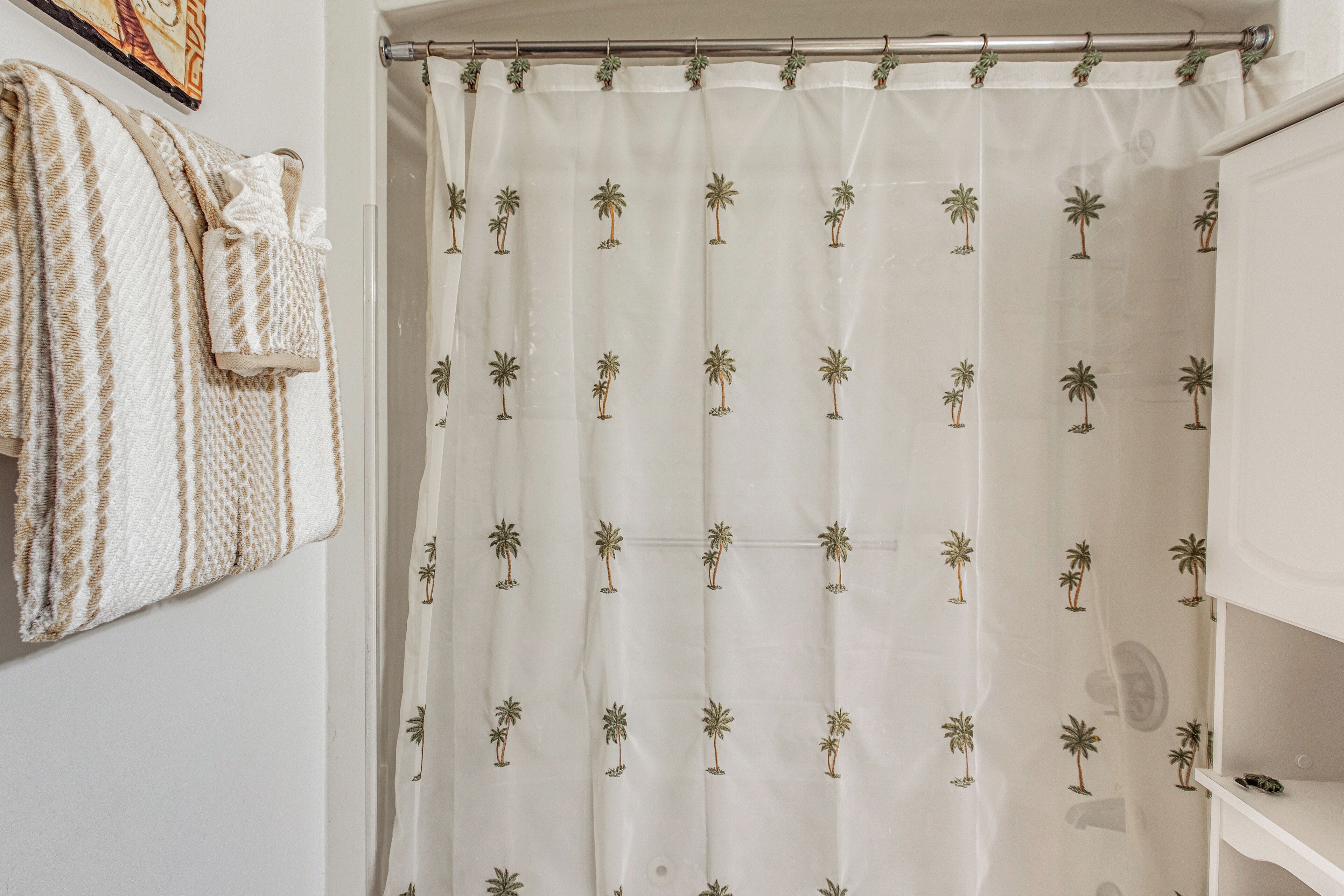A bathroom with a shower curtain featuring palm tree patterns, a towel rack with a patterned towel, and a white shelf with a small item on it.