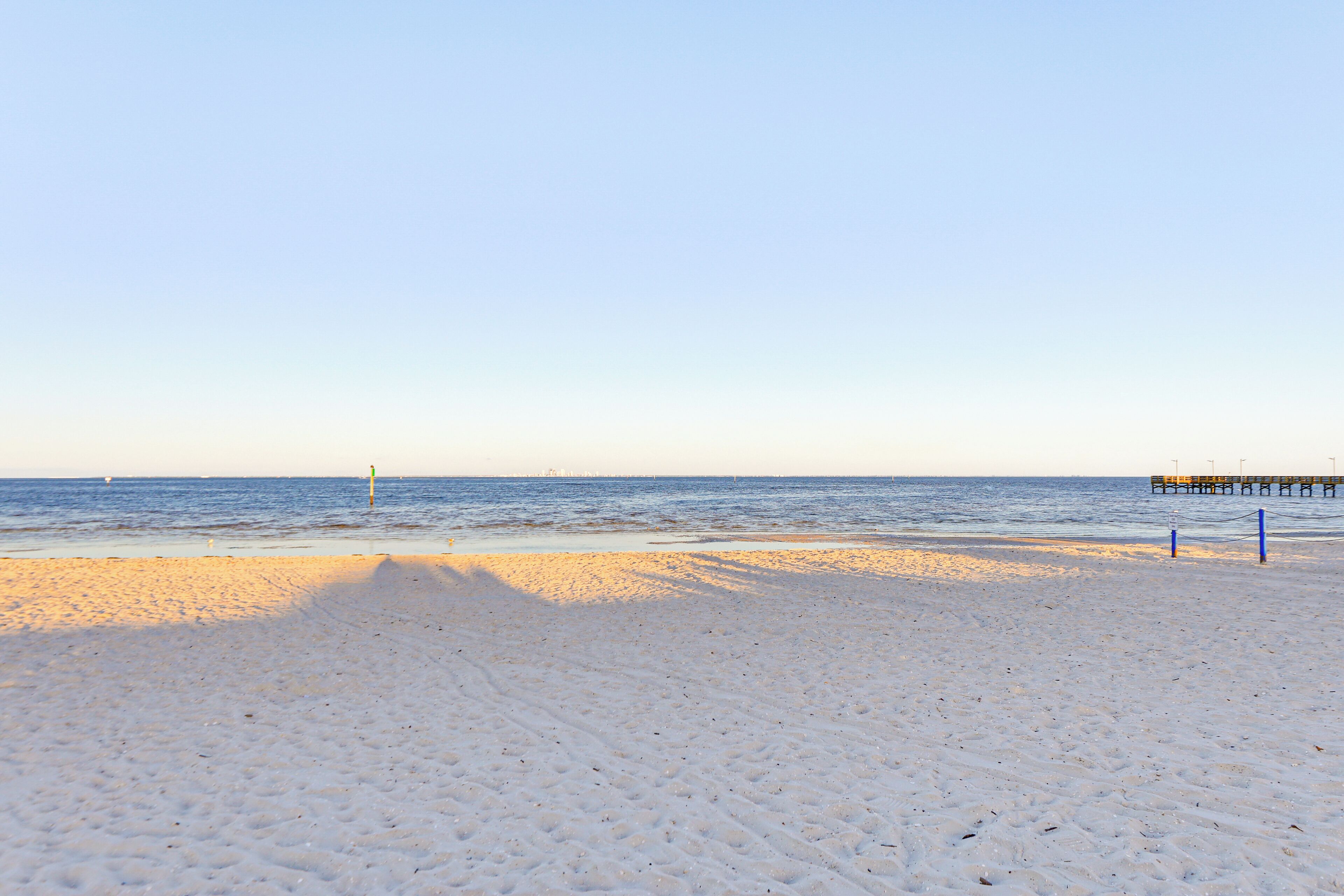 En strand med klar himmel, stilla hav och en pir i fjärran.