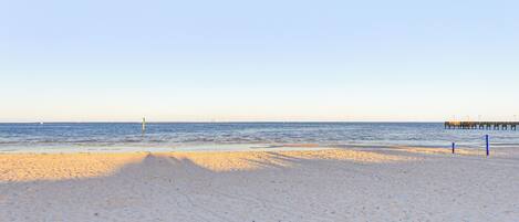 En strand med klar himmel, stilla hav och en pir i fjärran.