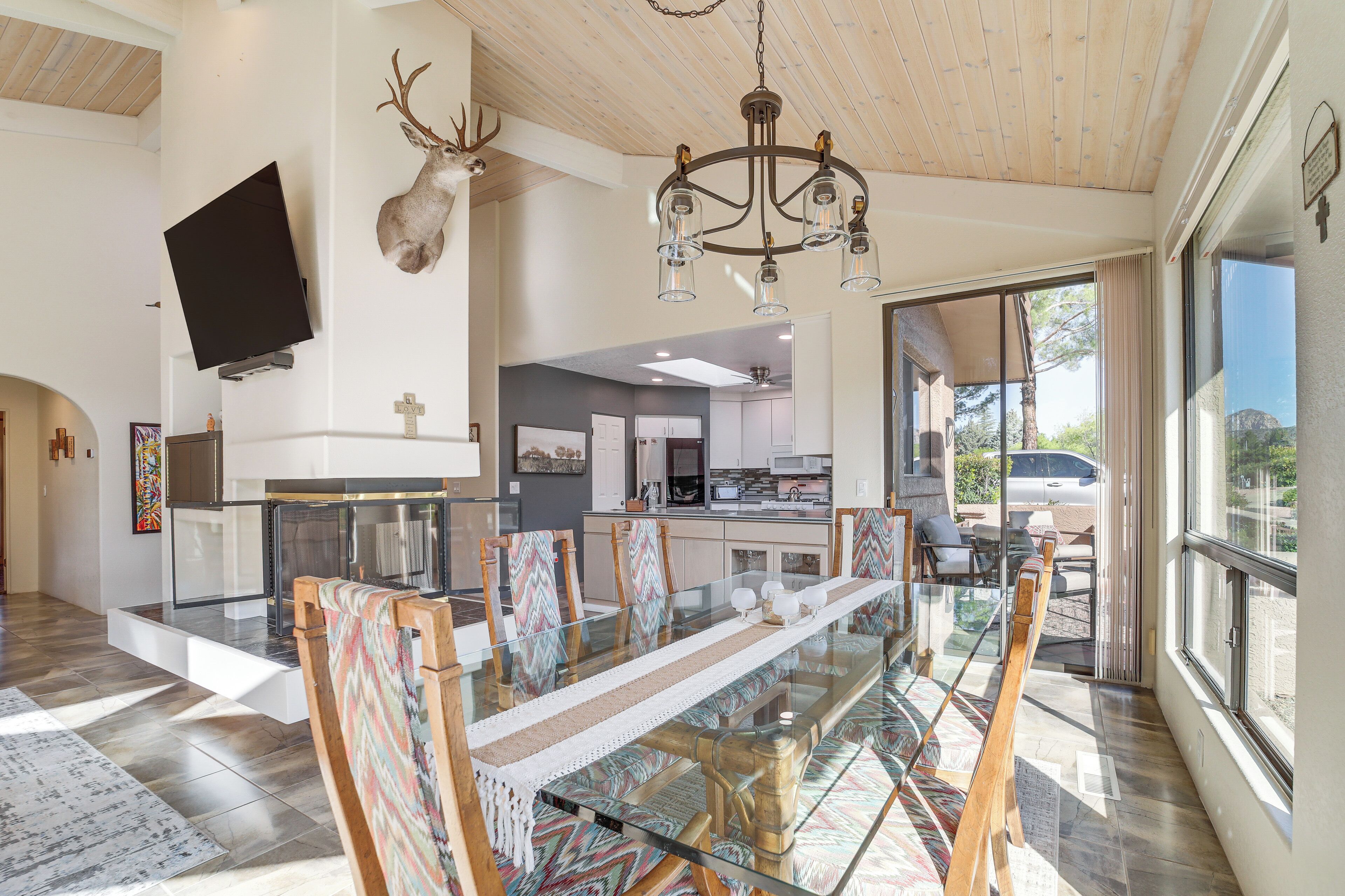 A spacious dining area with a glass table, wooden chairs, a chandelier, a mounted TV, and a deer head on the wall.