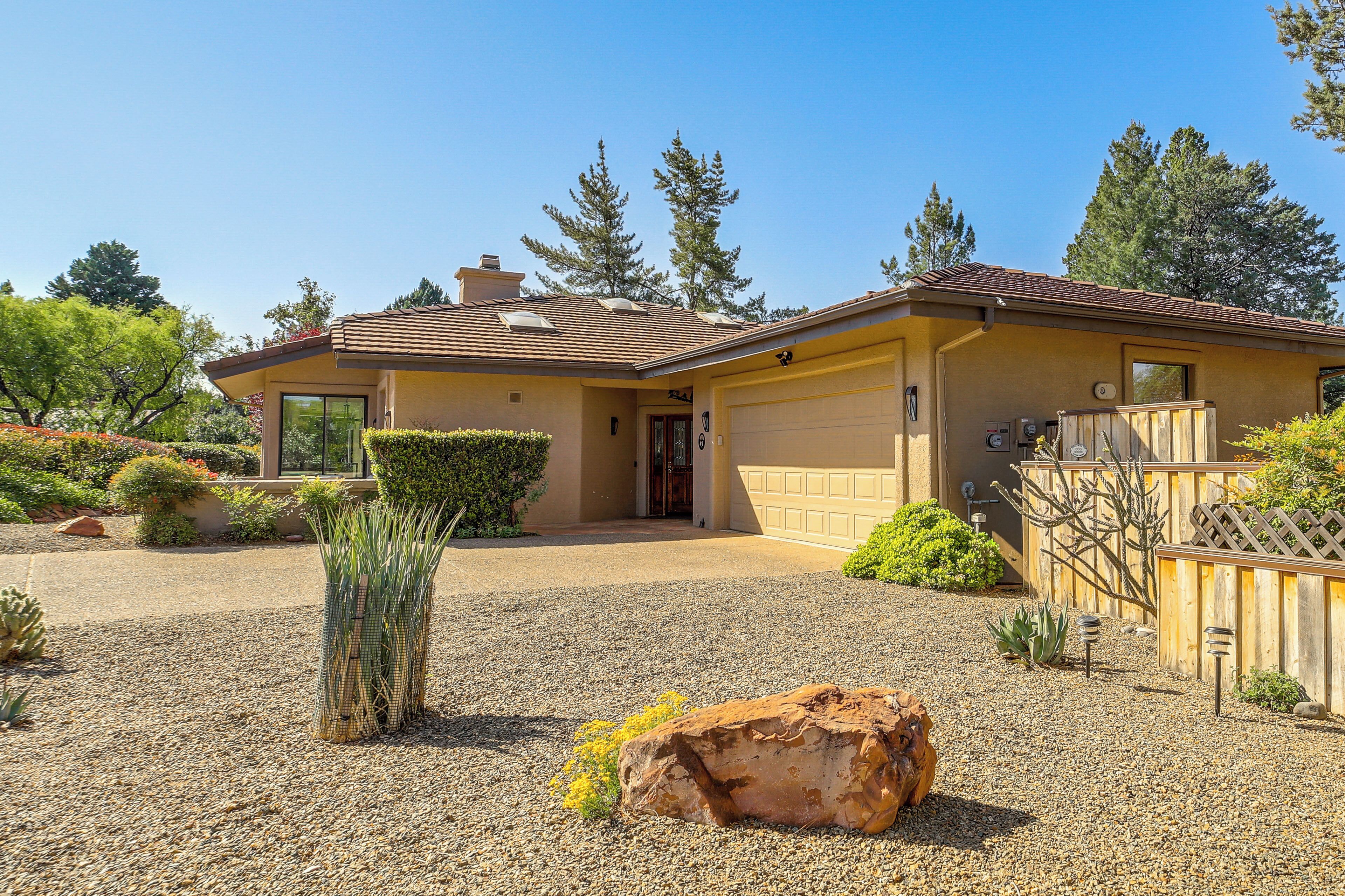 A single-story house with a garage, a gravel driveway, and a well-maintained front yard with various plants and a large rock.