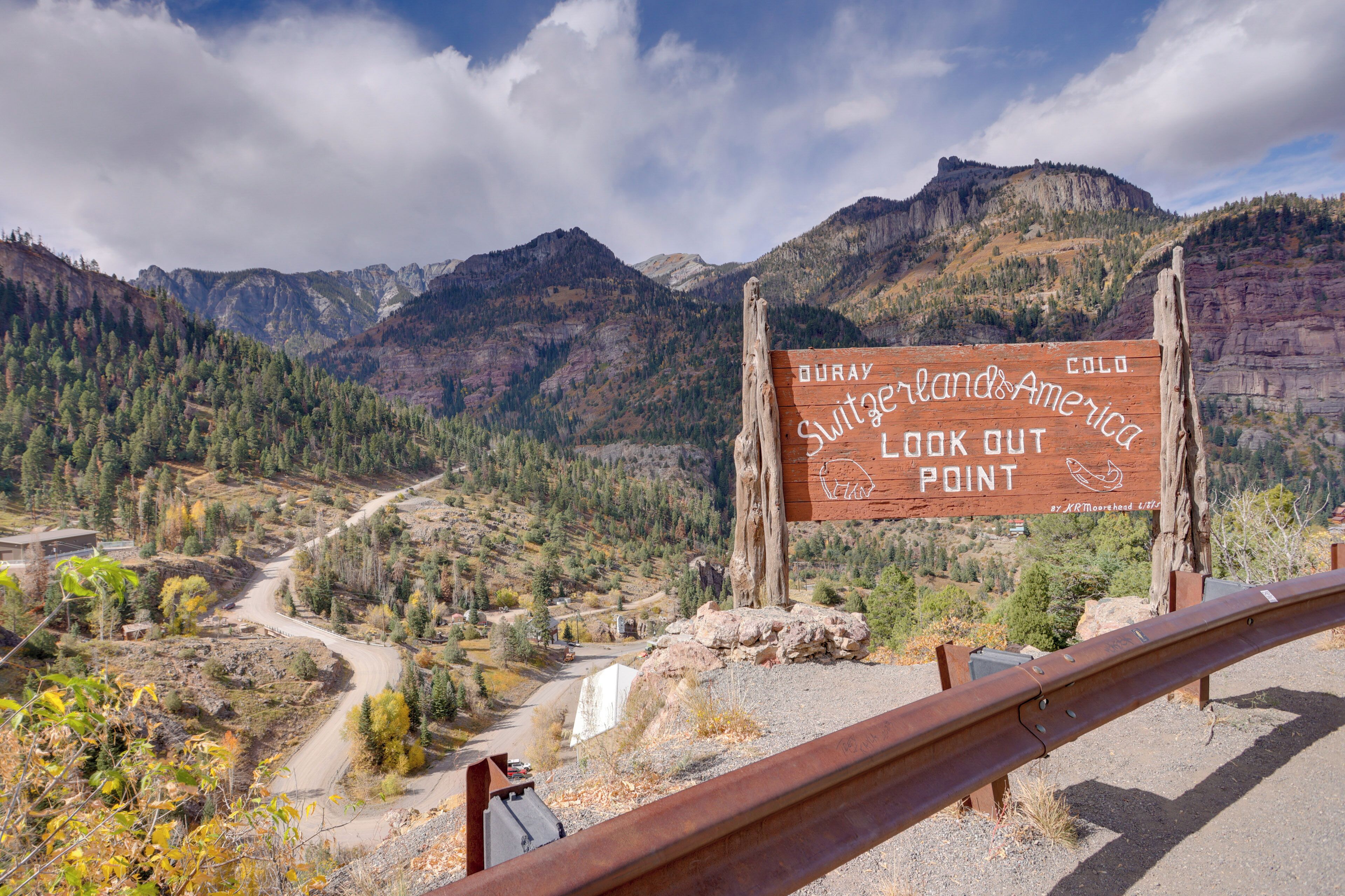 A wooden sign with text, a road, and mountainous terrain.