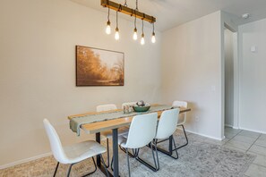 Un comedor con una mesa de madera, sillas blancas, una lámpara y un cuadro en la pared.