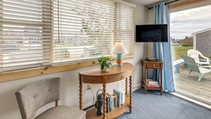 A room with a TV, a table with a lamp and books, a chair, and a view through a window with blinds.
