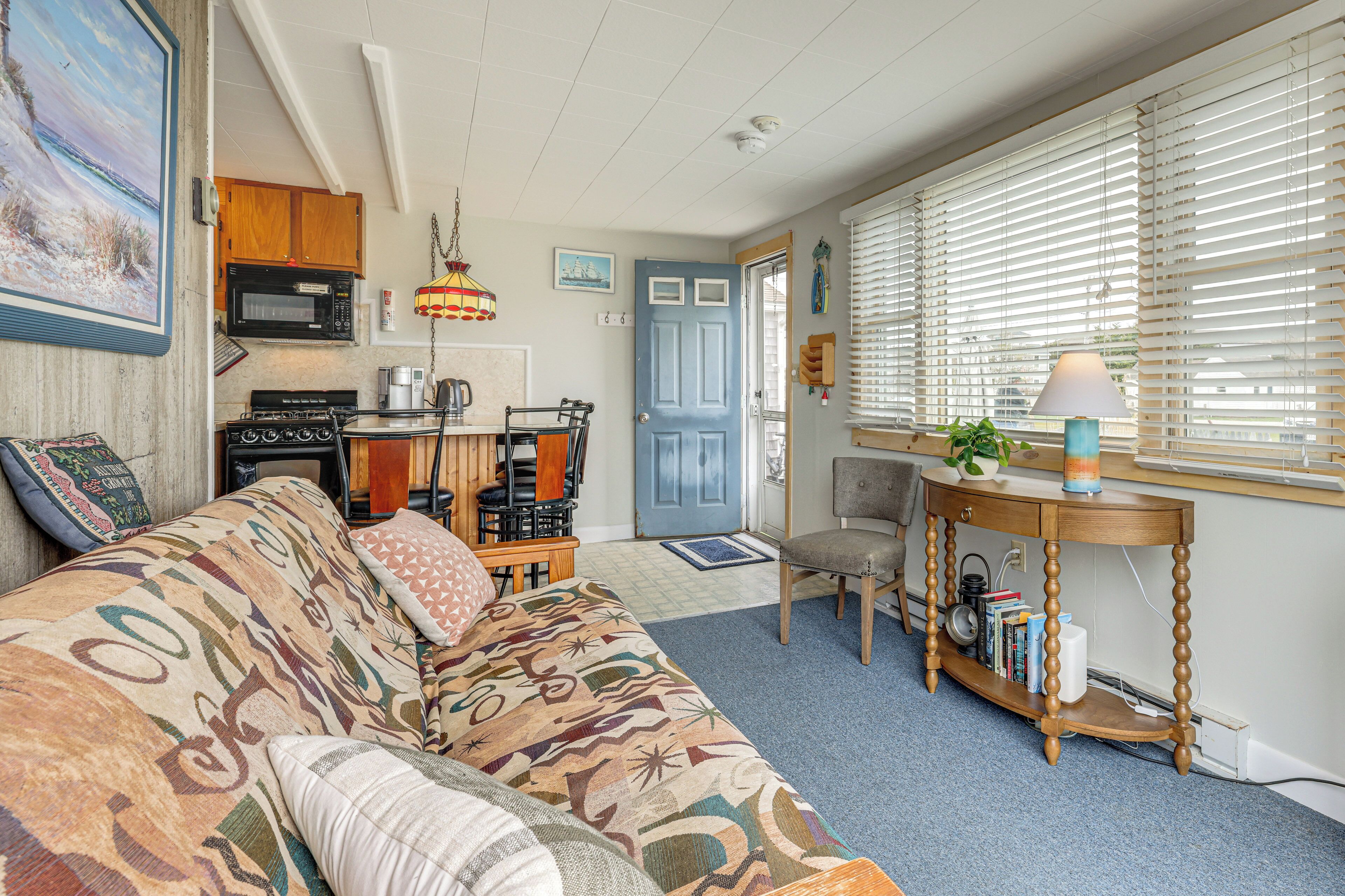 A cozy living room with a patterned sofa, a dining area with a table and chairs, a kitchen with appliances, and a window with blinds.