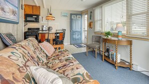 A cozy living room with a patterned sofa, a dining area with a table and chairs, a kitchen with appliances, and a window with blinds.