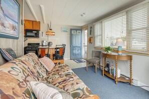 En hyggelig stue med en stribet sofa, et spiseområde med bord og stole, et køkken med hårde hvidevarer, og et vindue med persienner.