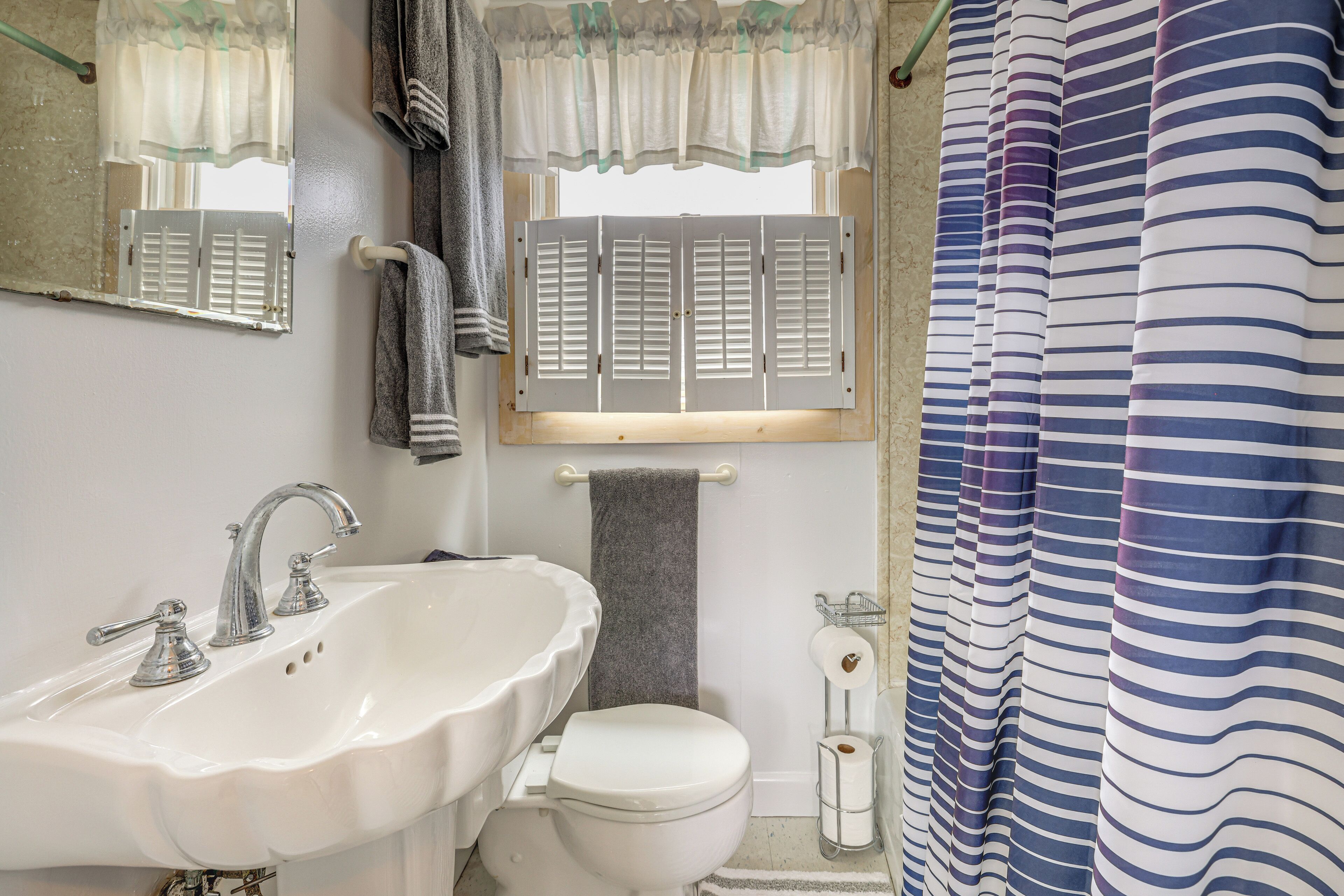 Un baño con un lavamanos blanco, dos grifos, un inodoro, un toallero, una ventana con contraventanas y una cortina de ducha a rayas.