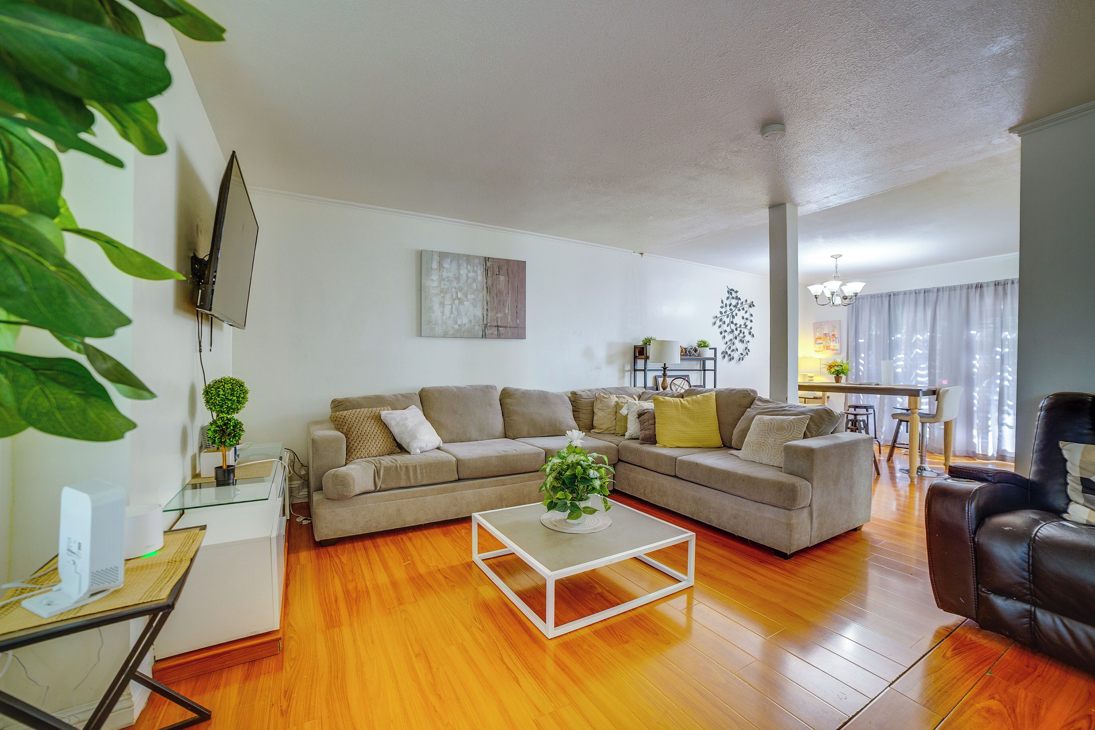 A living room with a sectional sofa, a glass coffee table, a TV mounted on the wall, and a dining area in the background.