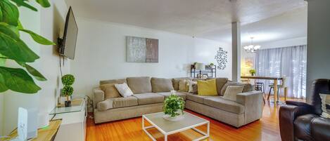 A living room with a sectional sofa, a glass coffee table, a TV mounted on the wall, and a dining area in the background.