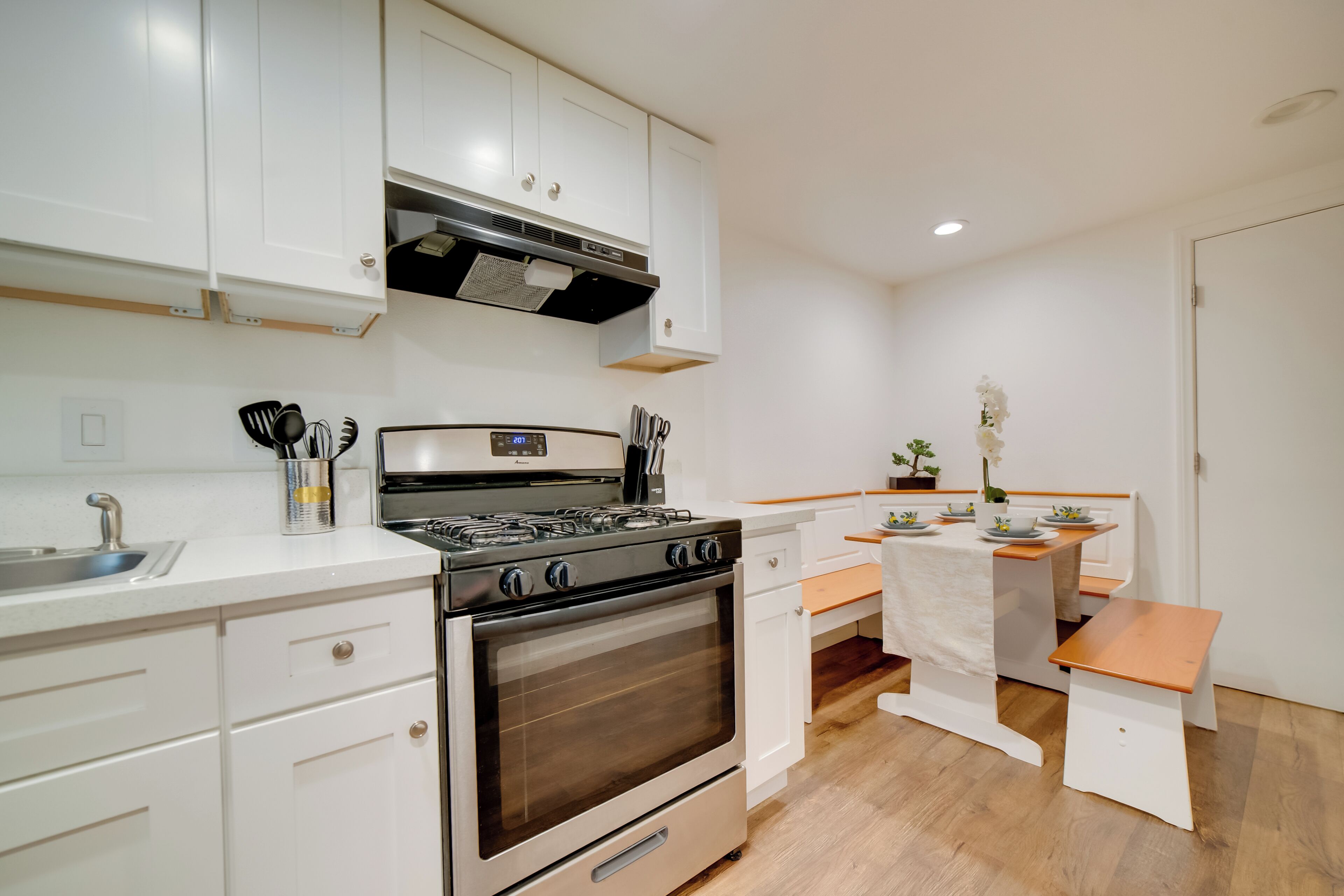 A kitchen with white cabinets, a stove, a sink, and a dining area with a bench.