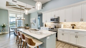 Una cocina moderna con armarios blancos, una isla de mármol, taburetes de bar y un amplio ventanal.
