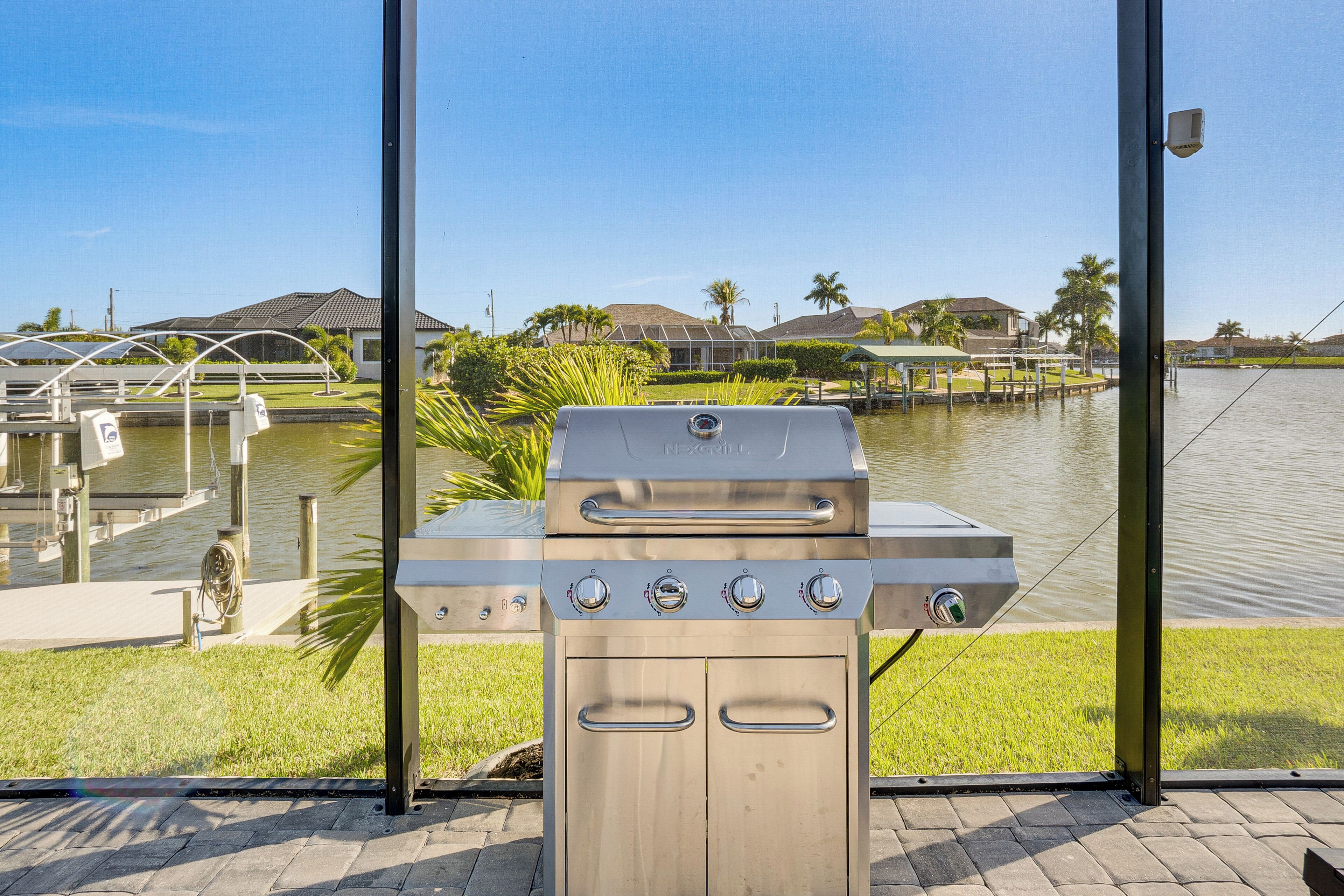 Un barbecue en acier inoxydable doté de quatre brûleurs, d’un brûleur latéral et d’un bac à eau, situé près d’une étendue d’eau, avec une jetée et des maisons en arrière-plan.
