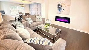 Ein Wohnzimmer mit einem Ecksofa, einem Glastisch, einem Kamin und einem Essbereich im Hintergrund.