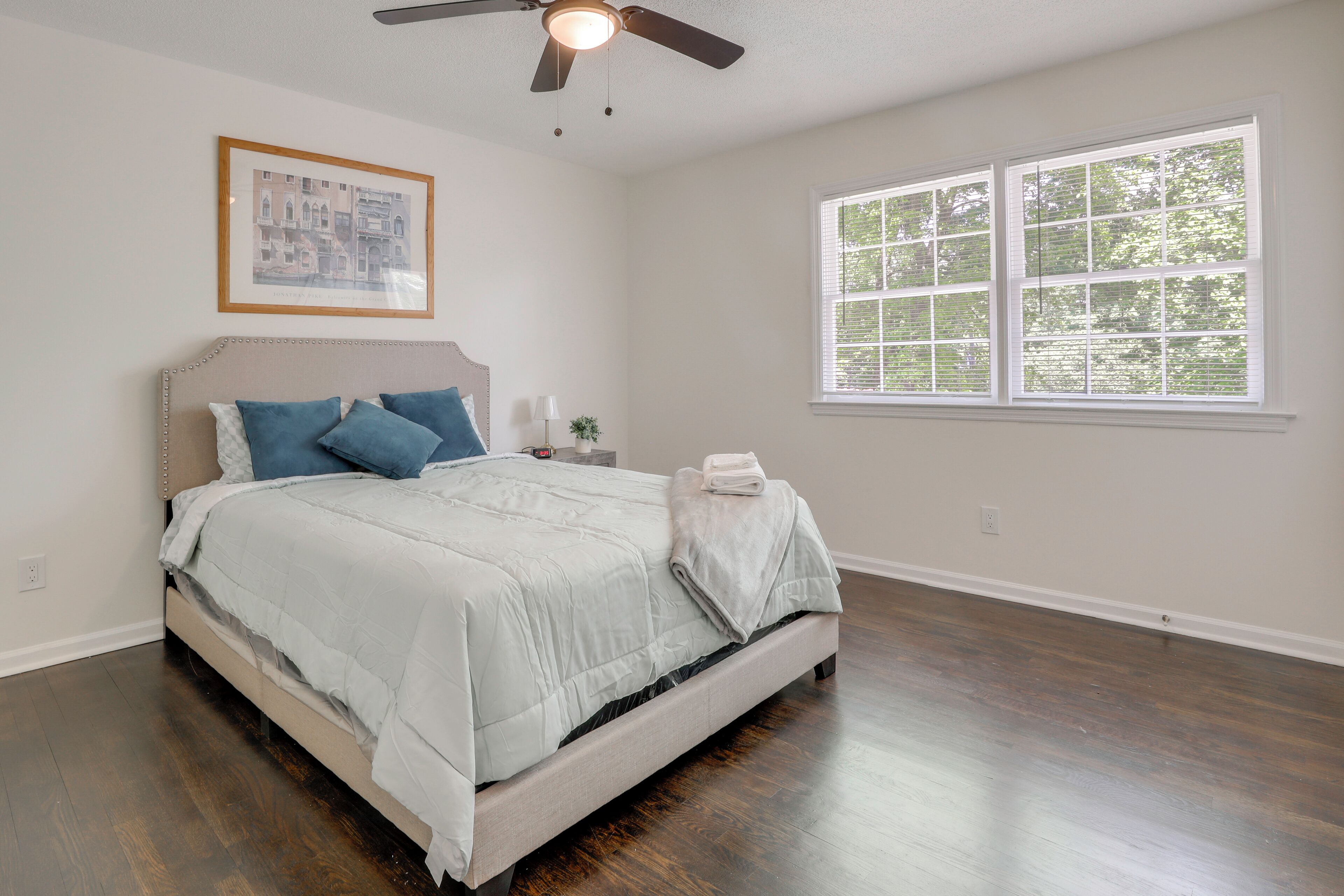 A bedroom with a bed, a ceiling fan, a window, and a framed picture on the wall.