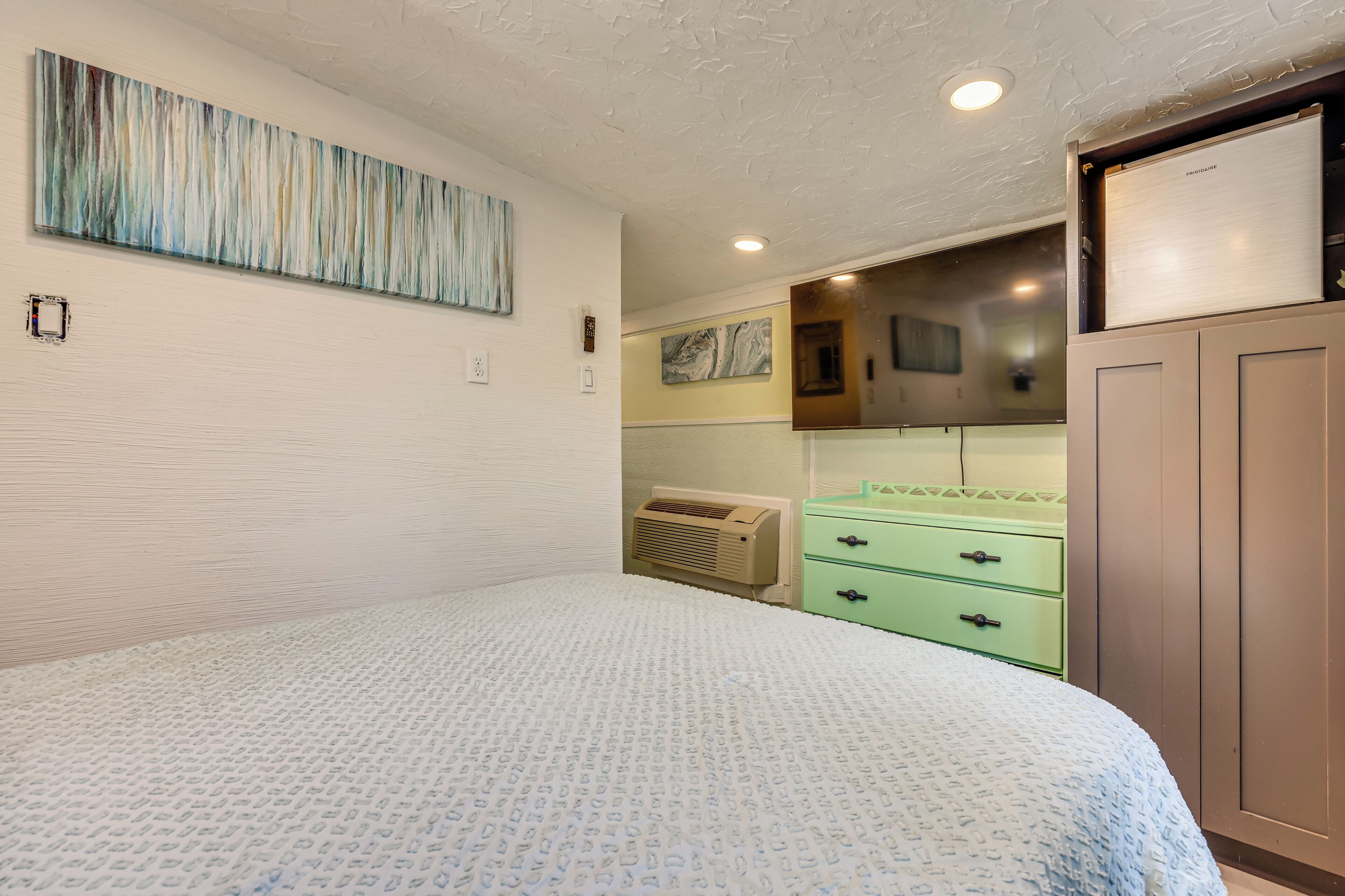 A bedroom with a bed, a green dresser, a wall-mounted air conditioner, and a painting.