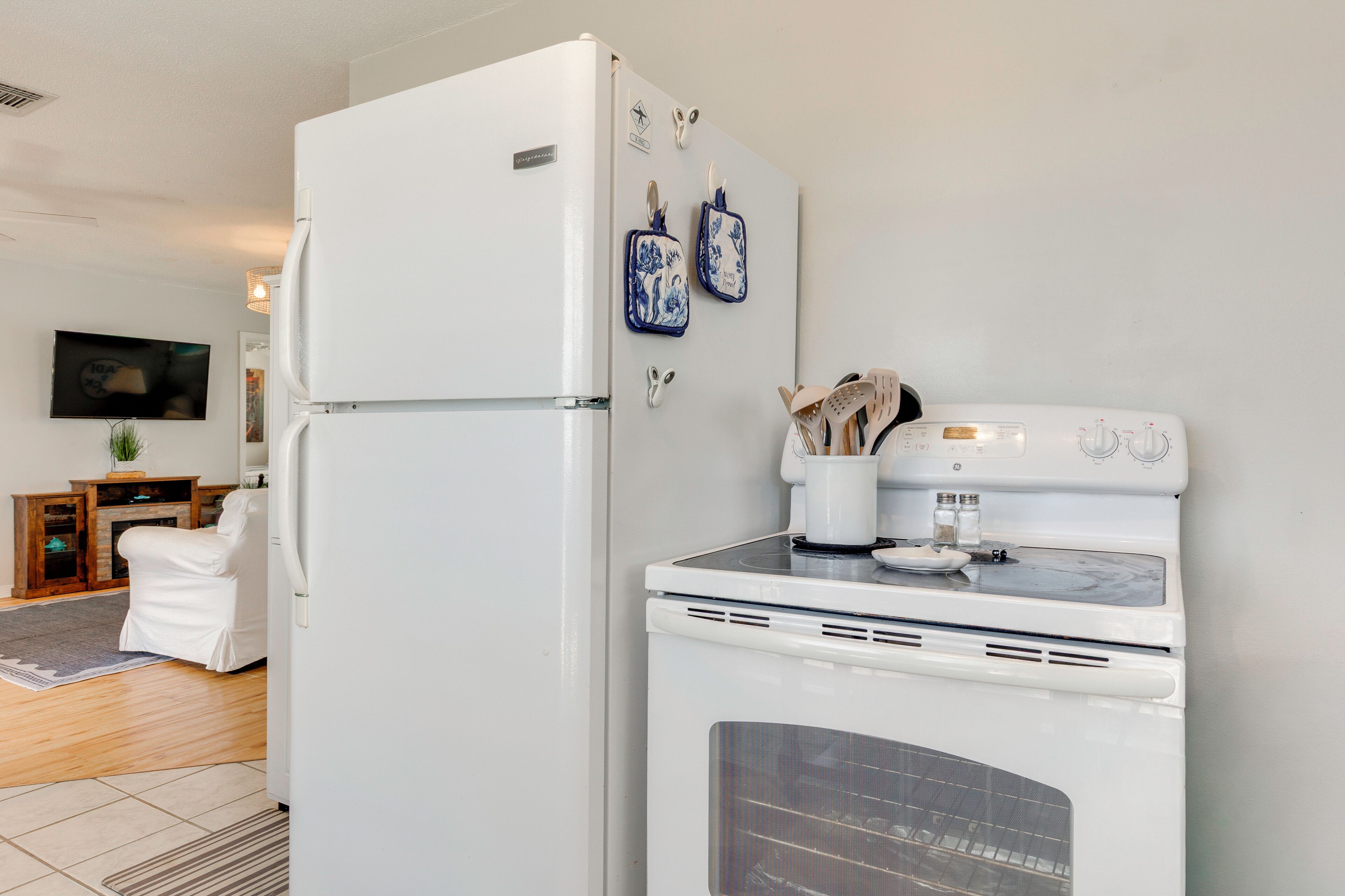 A kitchen with a refrigerator, oven, and microwave. There is a television on the wall and a white chair in the living area.