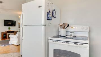 A kitchen with a refrigerator, oven, and microwave. There is a television on the wall and a white chair in the living area.