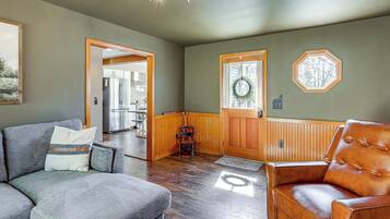 A living room with a grey couch, a brown leather chair, wooden flooring, a wooden door with a wreath, and a view into the kitchen.