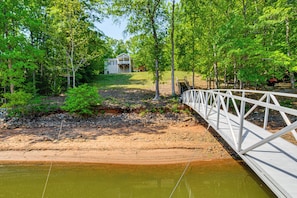 A wooden bridge over a river, surrounded by trees and a house in the background.