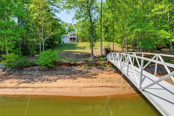 Eine Holzbrücke über einem Fluss, umgeben von Bäumen und ein Haus im Hintergrund.