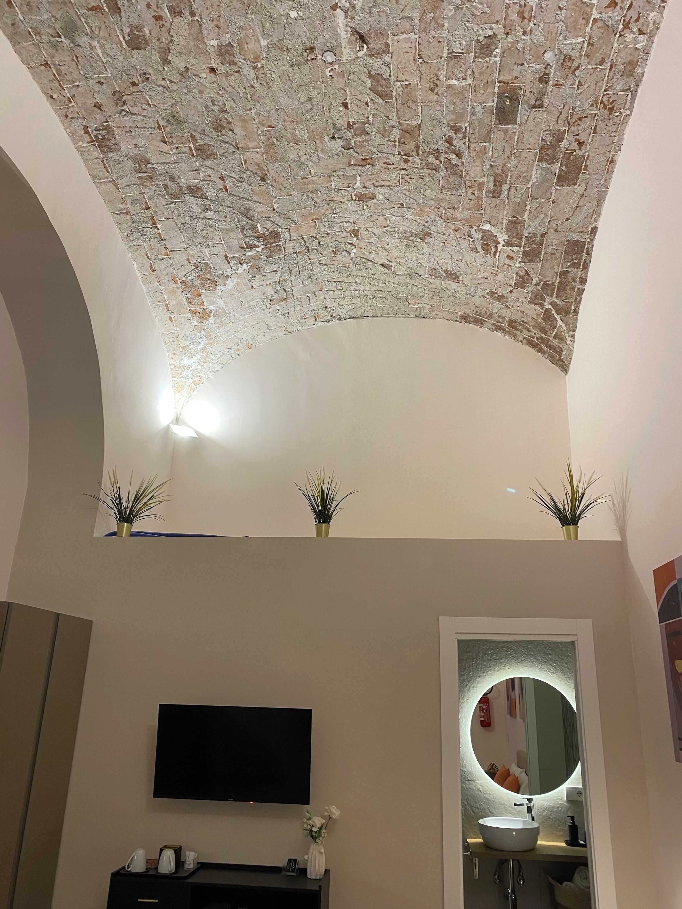 A room with a brick ceiling, a flat-screen TV, a round mirror, and a sink.