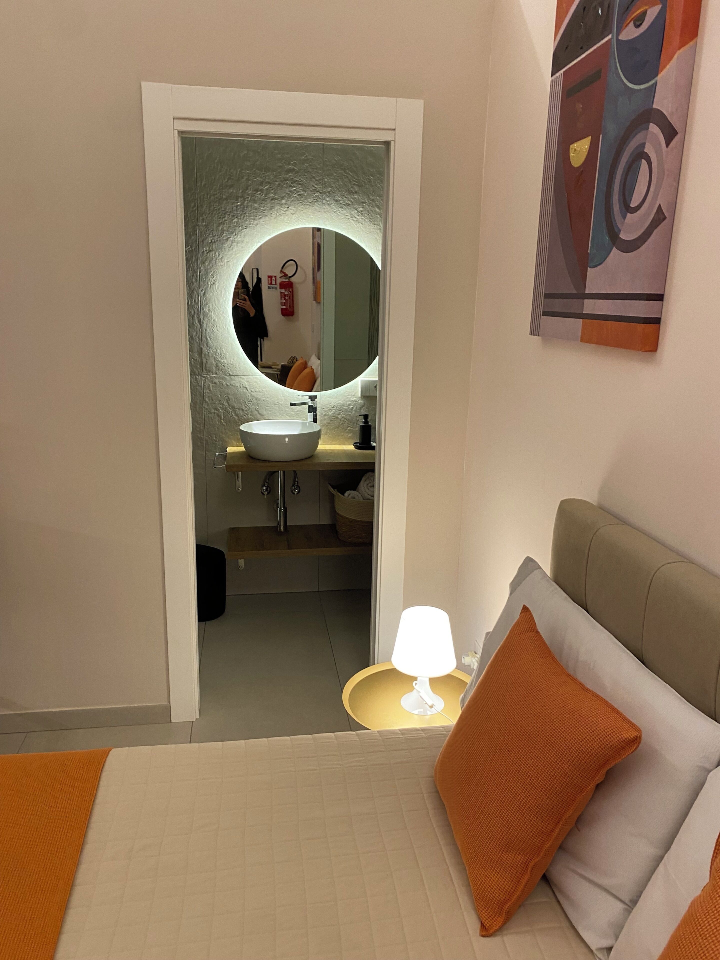 Une chambre d’hôtel avec un lit, une lampe de chevet, un miroir rond, un lavabo et un tableau au mur.