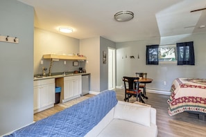 Quarto compacto com cozinha compacta, uma cama com colcha azul, mesa de jantar com cadeiras e janela com cortinas.