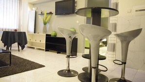 A modern living room with a TV mounted on the wall, a black sofa, a coffee table, and three bar stools. - Fana Hotel (Dakar)