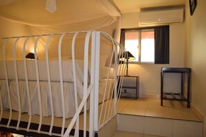 A single bed with a white metal frame, a bedside table with a lamp, a window with curtains, and a wall-mounted air conditioner. - Fana Hotel (Dakar)