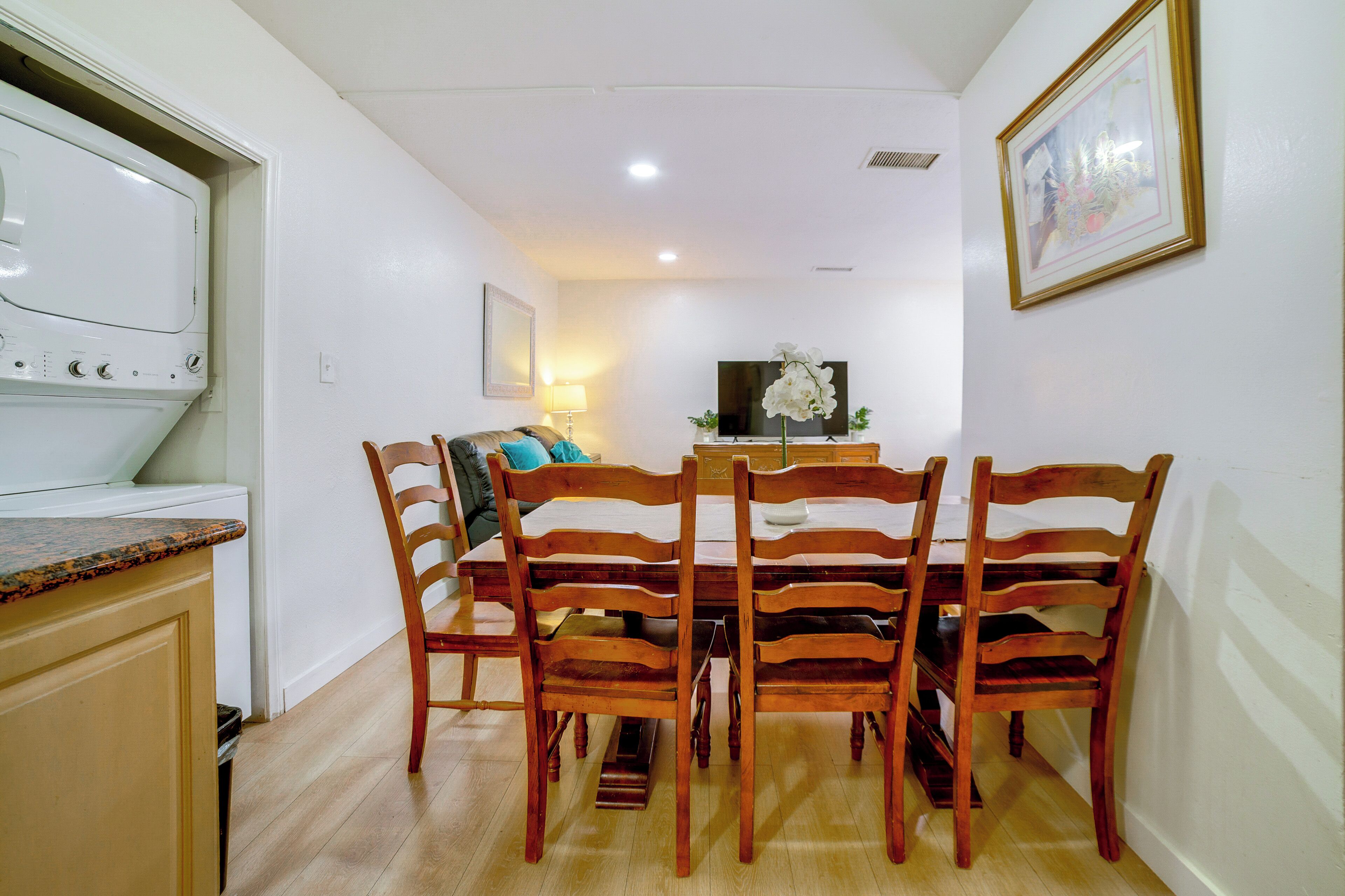 Uma sala de jantar com mesa e cadeiras de madeira, uma televisão e uma cozinha com fogão.