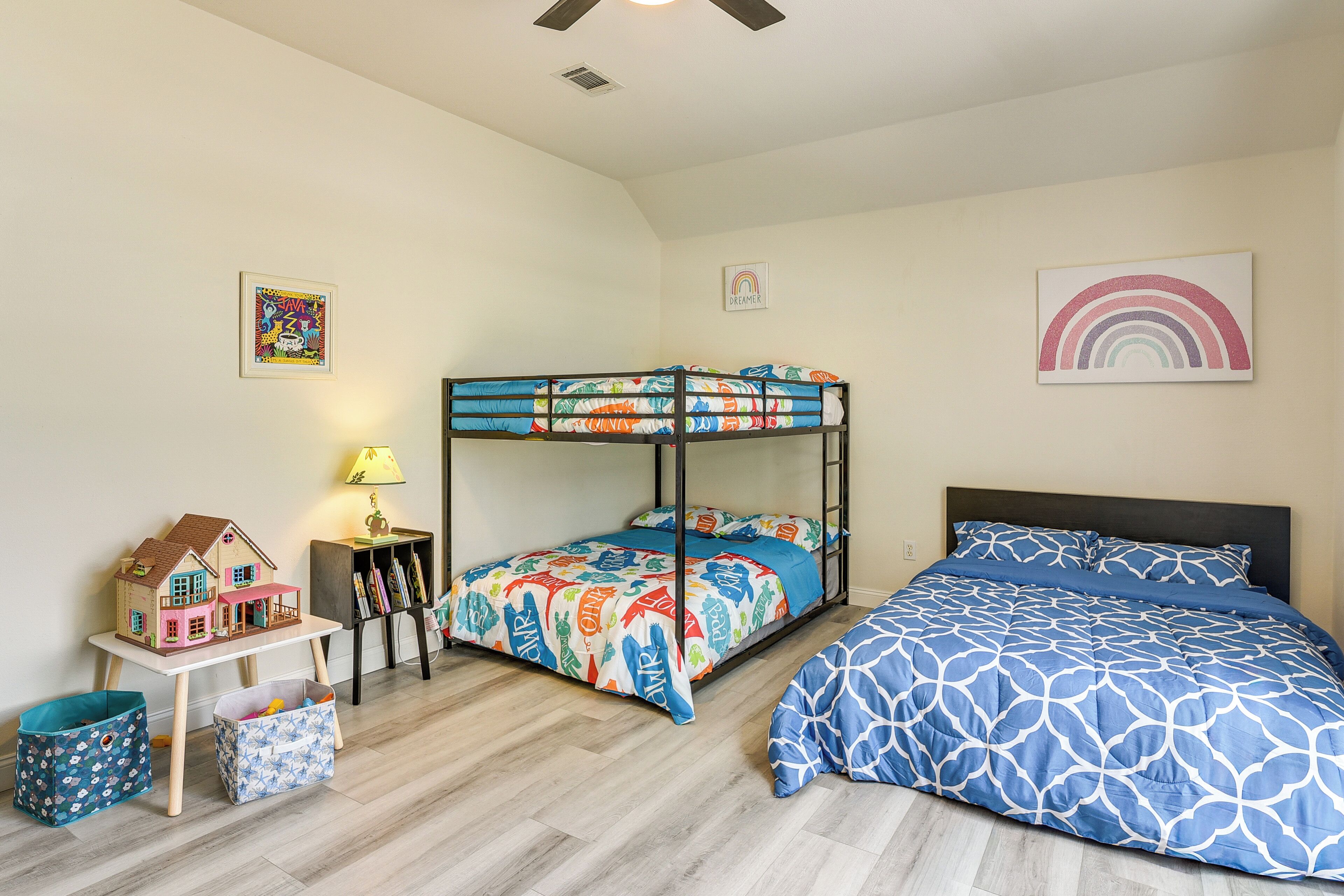Una habitación con literas, una pequeña mesa con una lámpara, una casita de juguete y un cuadro de un arcoíris en la pared.