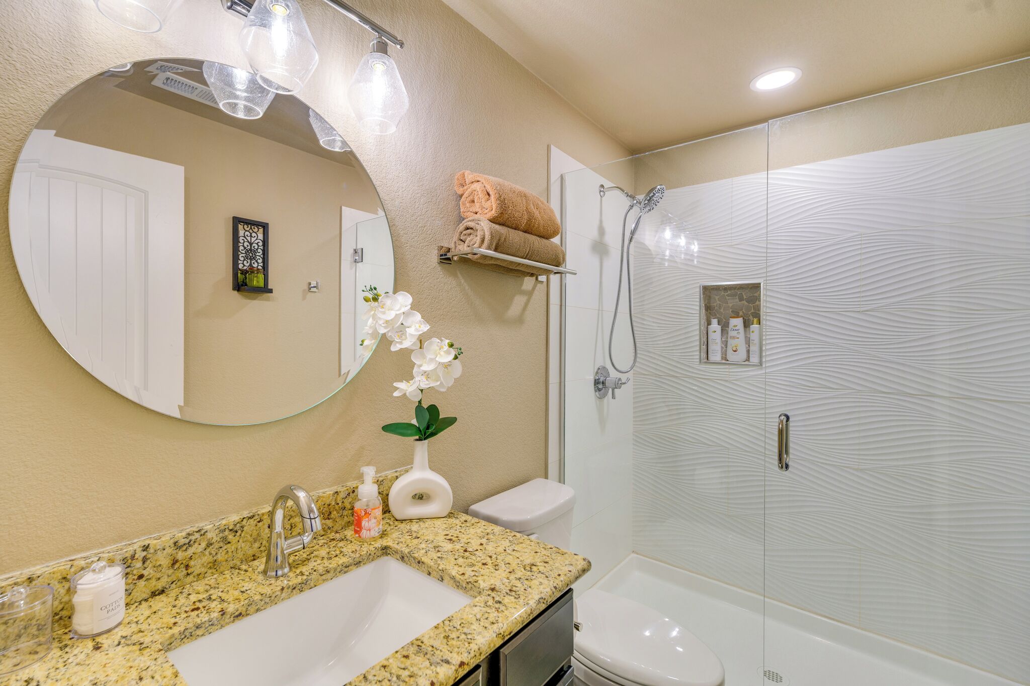 A bathroom with a granite countertop, a round mirror, a white sink, a toilet, a shower with a glass door, and a towel rack with towels.