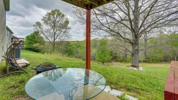 A glass table under a wooden pergola with a view of a grassy yard and trees.