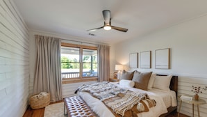 A bedroom with a bed, a bench, a ceiling fan, a window with curtains, and framed pictures on the wall.