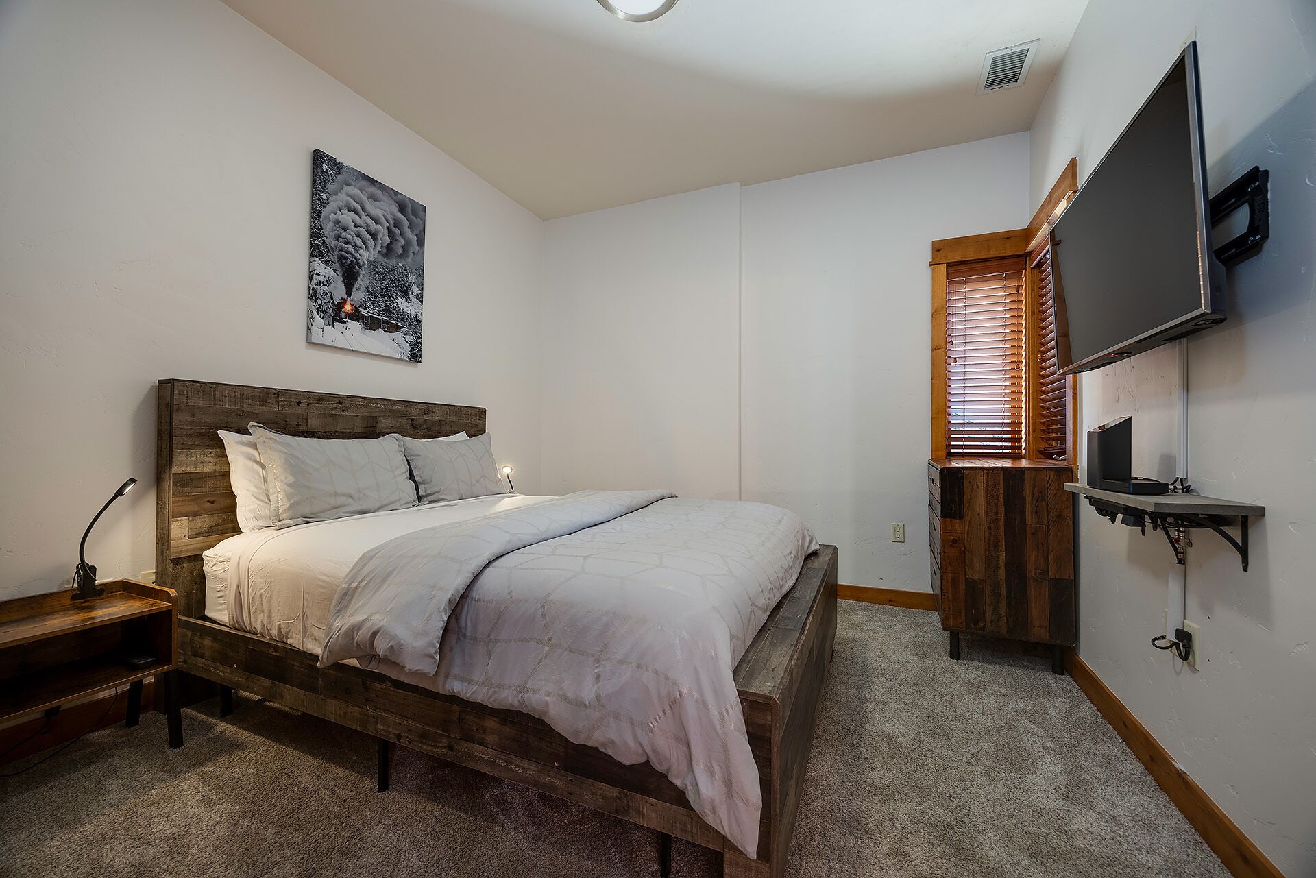 A bedroom with a wooden bed, a nightstand, a TV mounted on the wall, and a window with blinds.