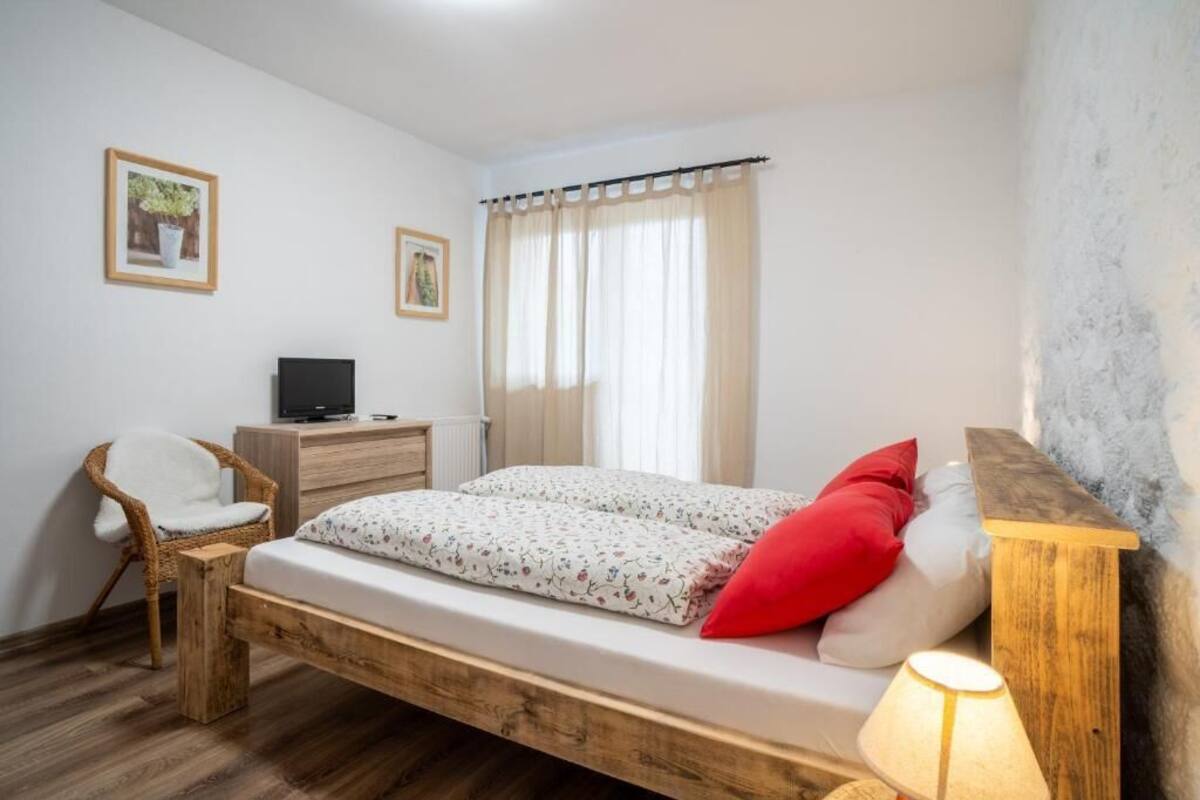 A bedroom with a wooden bed, a chair, a desk with a TV, and a window with curtains.