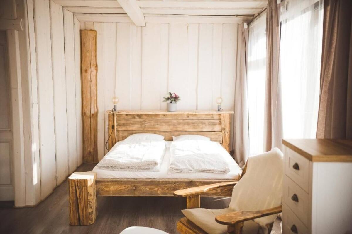 A wooden bed with a headboard, a bedside table, a chair, and a dresser in a room with wooden walls and a window.