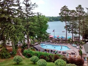 A pool area with lounge chairs and umbrellas, surrounded by trees and a view of a lake with boats.