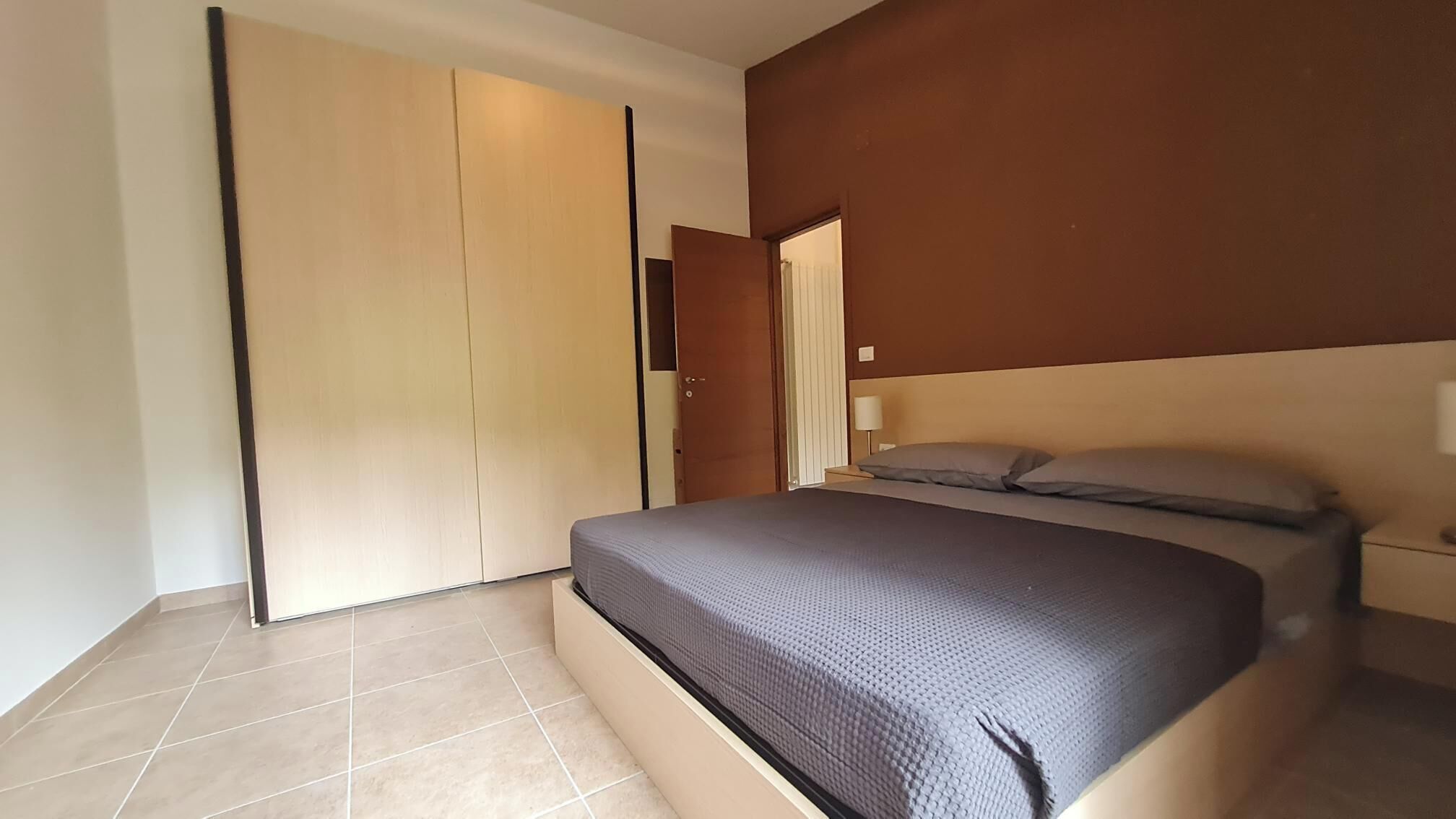 Ein Schlafzimmer mit Bett, Nachttisch, Lampe, Kleiderschrank und einer Tür.