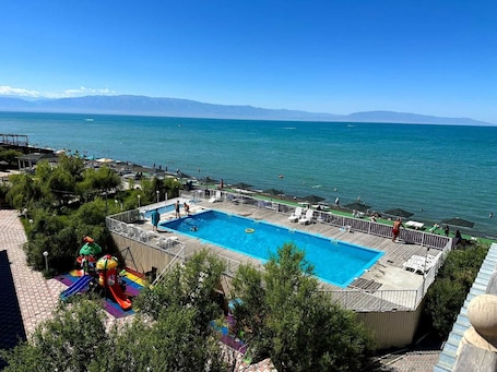 A swimming pool with a deck, surrounded by greenery, and overlooking a body of water with mountains in the distance.. Inju Alakol