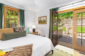 Un dormitorio con una cama, una mesita de noche con lámpara, un cuadro en la pared y vista a un jardín a través de un ventanal.