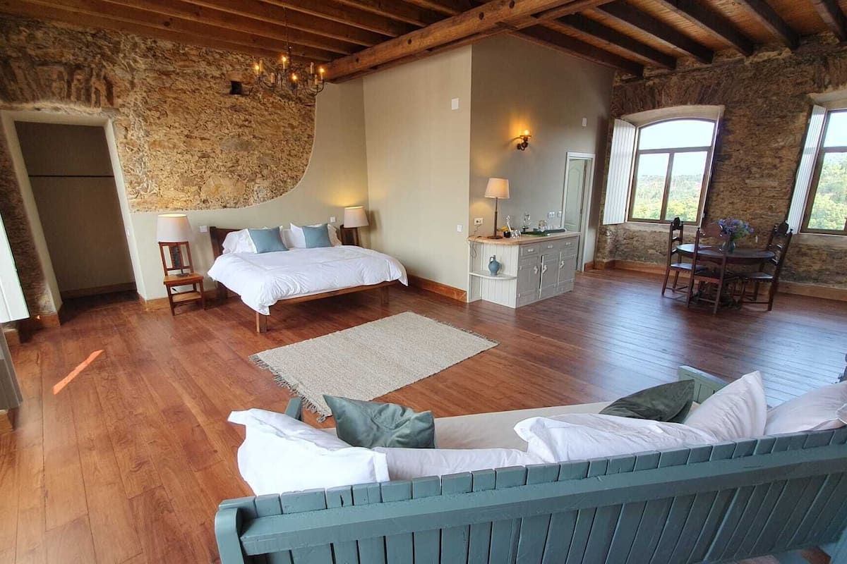 A bedroom with a large bed, a sitting area with a sofa, a small kitchen, and a stone wall.