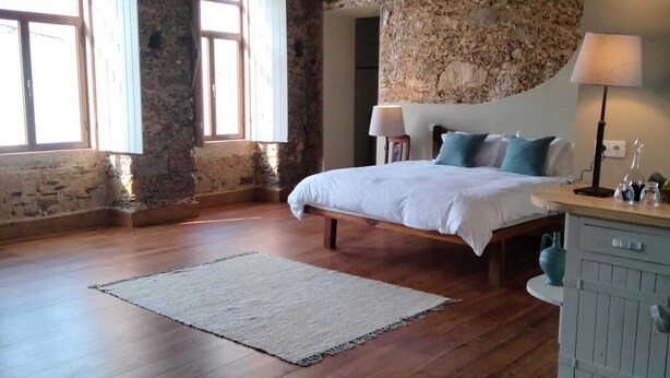 Une chambre avec un plancher en bois, un lit recouvert de linge de lit blanc, un mur en pierre et un lustre.