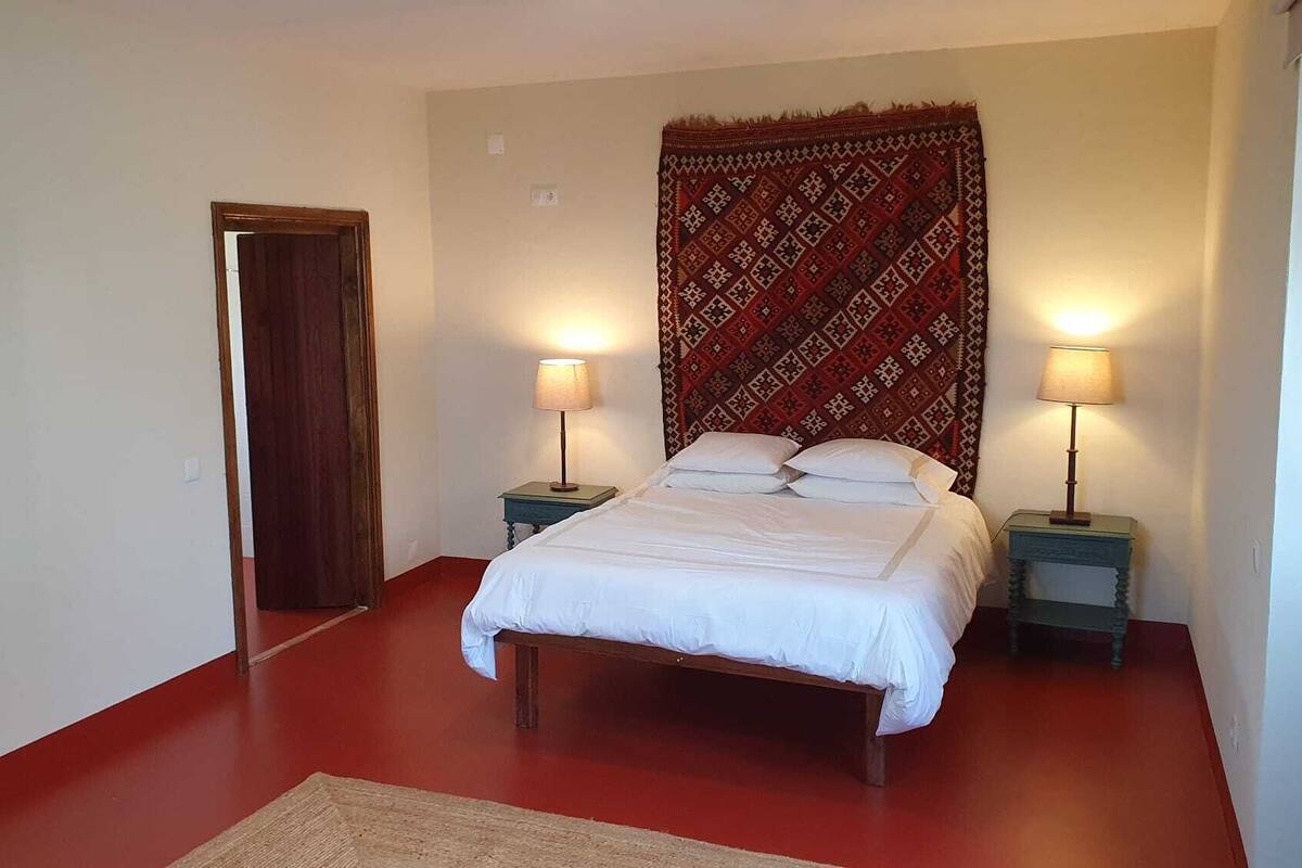 A bedroom with a bed, two bedside tables with lamps, a patterned tapestry, and a wooden door.