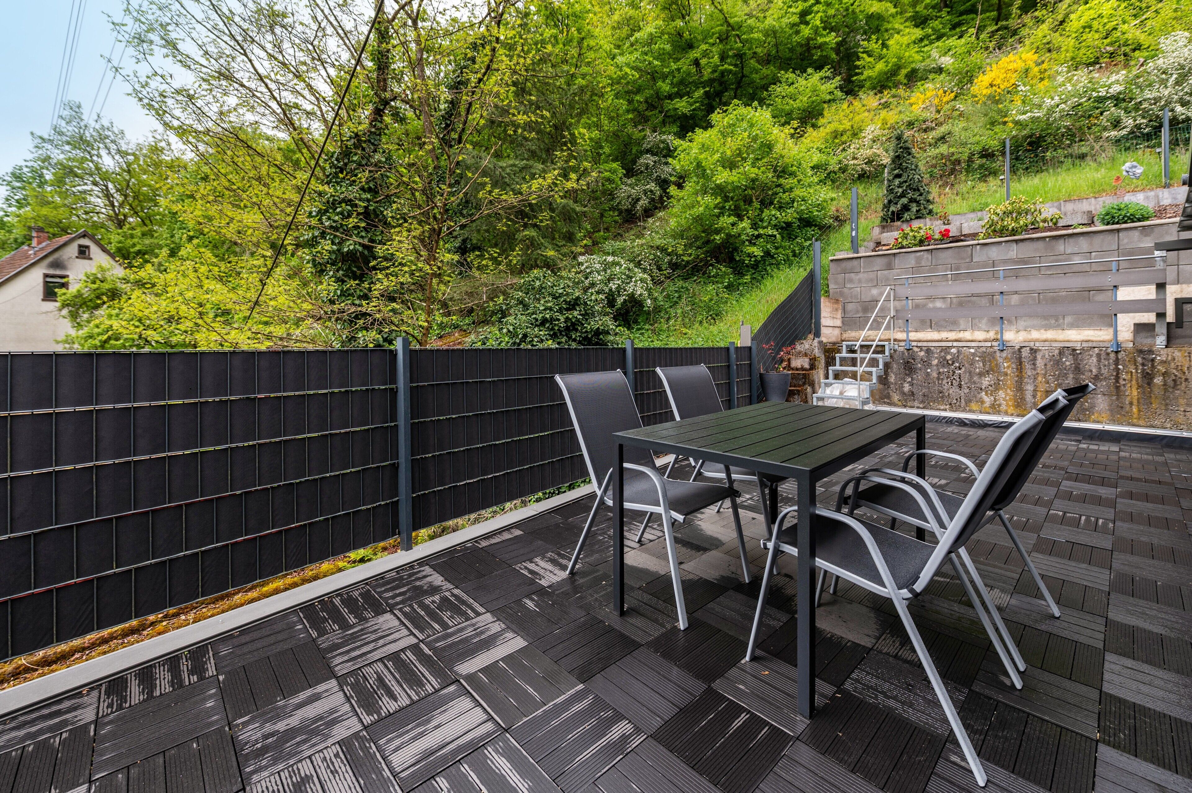 A patio with a table and chairs, a black fence, and a view of trees and a house.