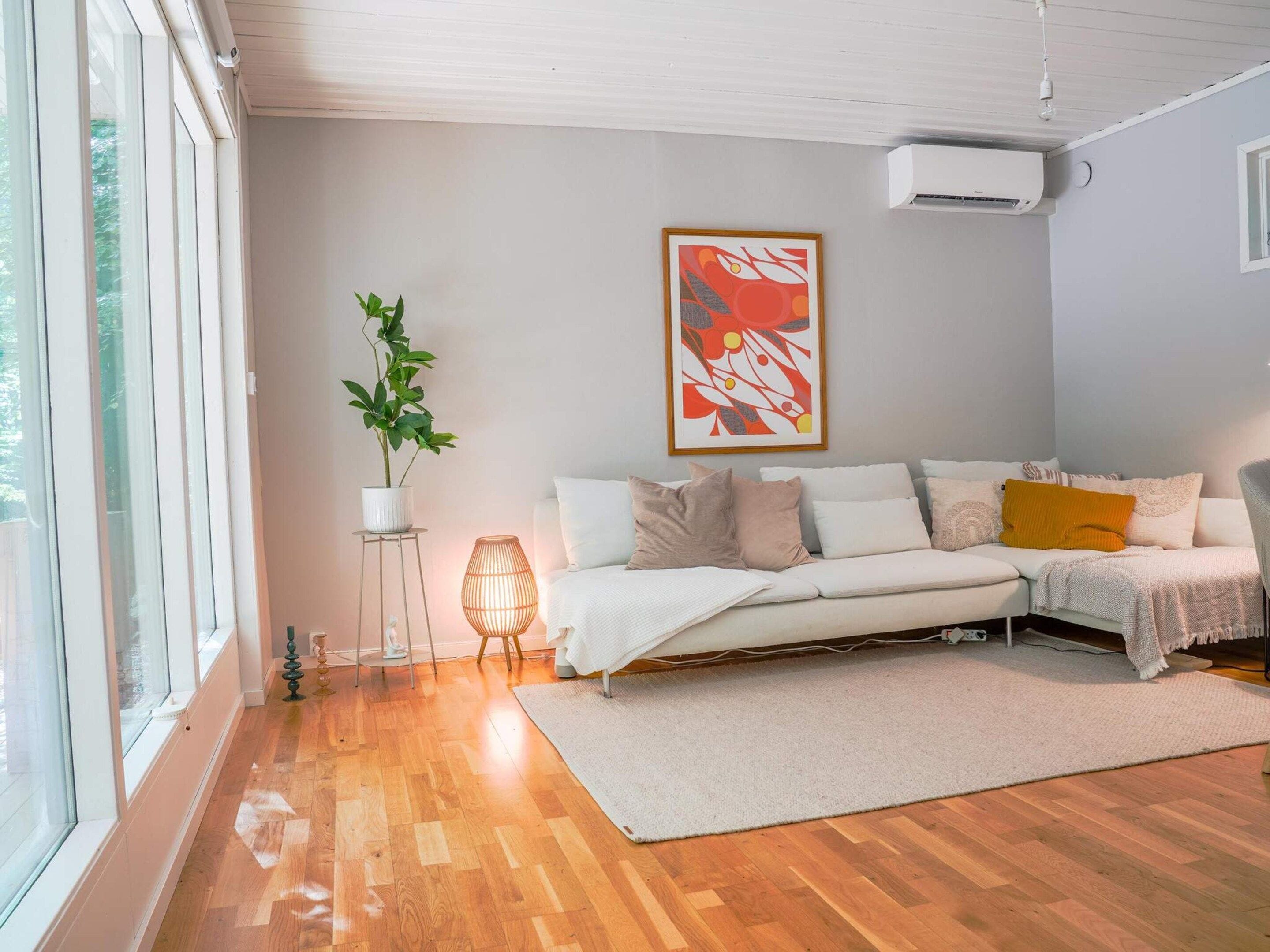 Una sala de estar con sofá, planta en maceta, lámpara de pie y aire acondicionado instalado en la pared.