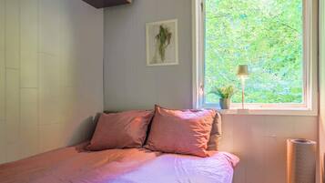 Un dormitorio con cama, ventana con vista a follaje, radiador y lámpara.