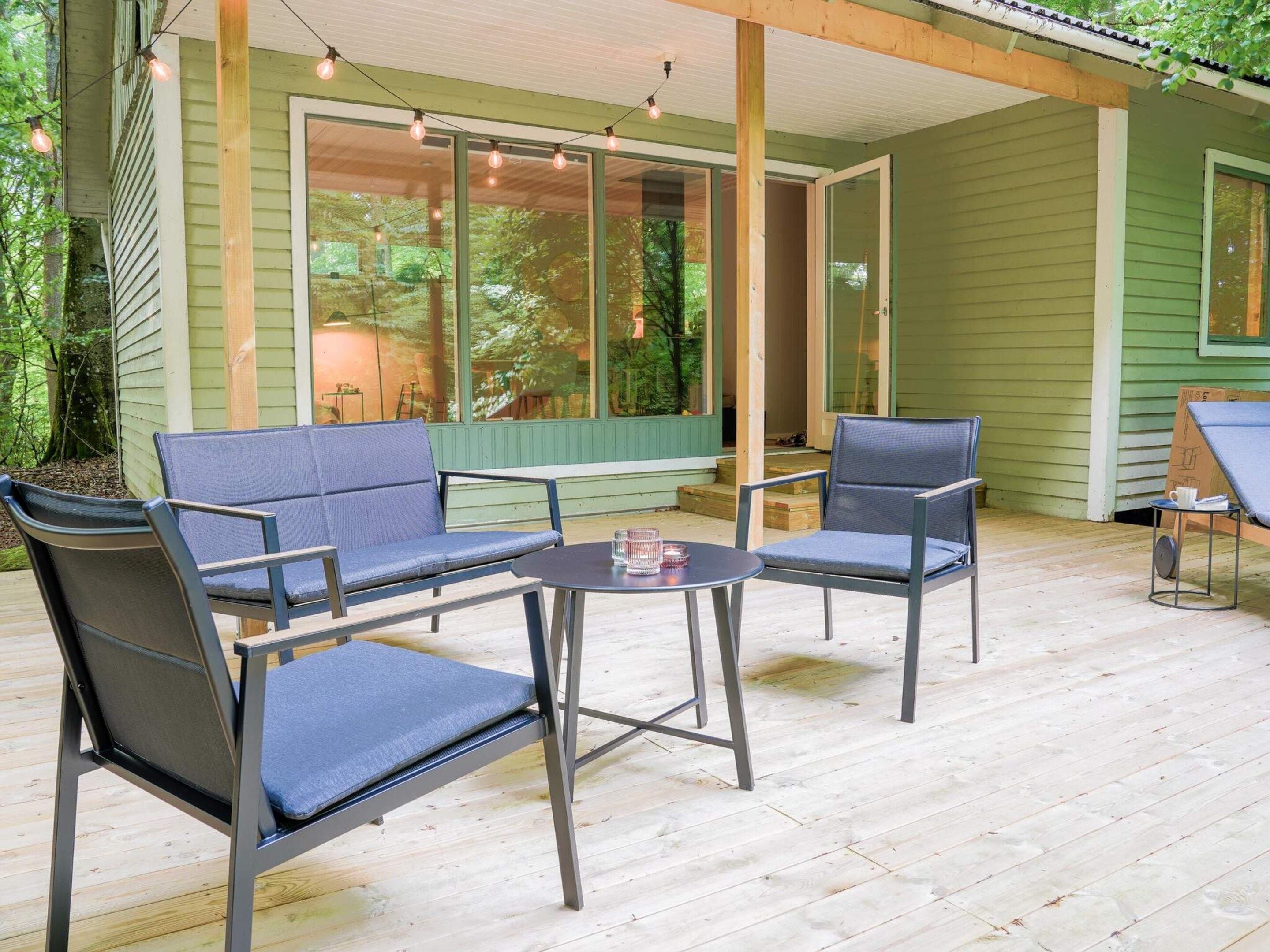 Un patio exterior con dos sillas y una mesa, un área cubierta con luces colgantes, y vista a una casa verde con ventanas grandes.